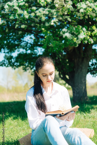 girl in a white blouse and blue jeans. Sitting near a flowering tree and reading a book.
