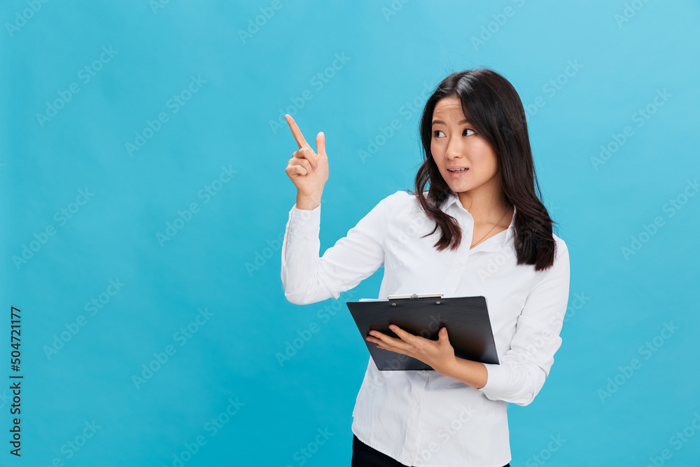 Excited cute Asian businesswoman in classic office dress code holds folder tablet with resume point finger up posing isolated on over blue studio background. Cool business offer. Job interview concept Excited cute Asian businesswoman in classic office dress code holds folder tablet with resume point finger up posing isolated on over blue studio background. Cool business offer. Job interview concept