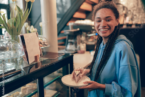 Cheerful stylsih mixed-race female bartender holding plate with piece of cake at counter. Portrait. photo