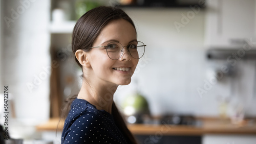 Happy young millennial woman wearing glasses posing in home kitchen banner head shot portrait. Positive nerdy smart student girl in stylish eyewear looking at camera, smiling photo