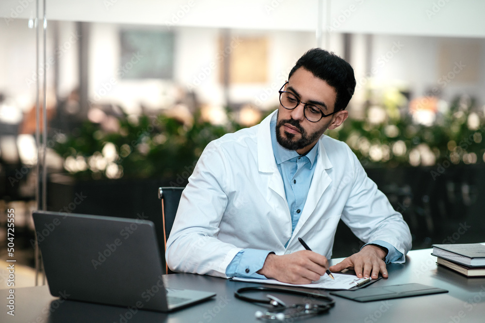 Obraz premium Serious attractive millennial arab guy doctor with beard in glasses, white coat looks at laptop, makes notes