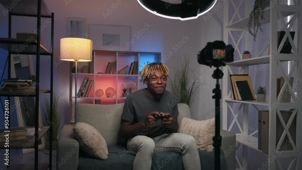 Gamepad review. Tech vlog. Excited male content creator sharing feedback on remote joystick controller for video game filming live stream on camera on tripod in modern home interior.