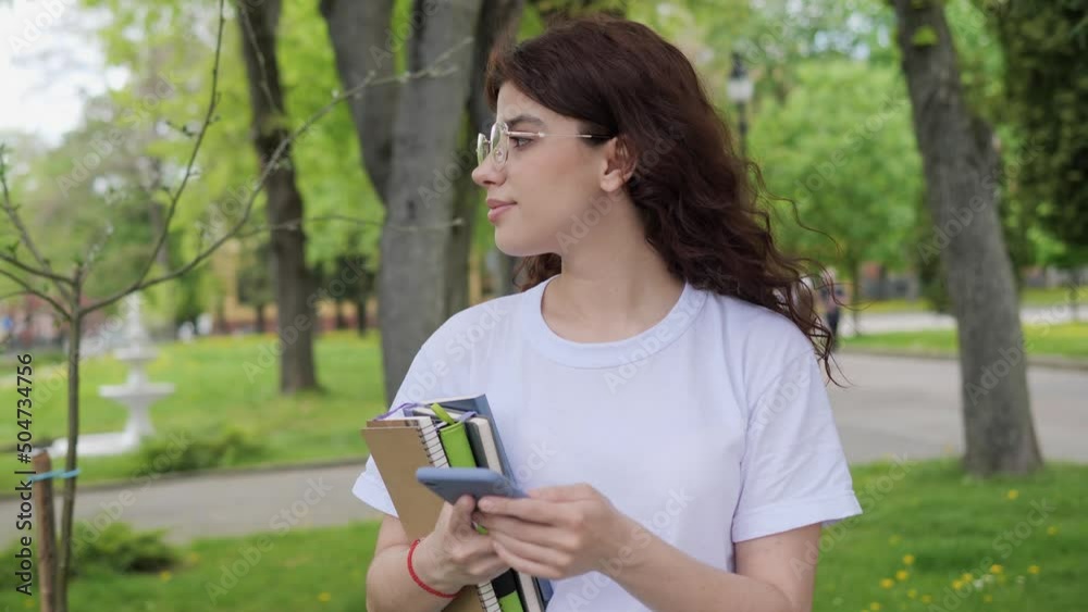 Beautiful young curly hair high school girl wearing white t-shirt and glasses messaging on smartphone surfing internet. Student holding notebooks with mobile phone standing in campus outdoors. Beautiful young curly hair high school girl wearing white t-shirt and glasses messaging on smartphone surfing internet. Student holding notebooks with mobile phone standing in campus outdoors.