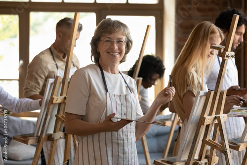 Mature woman painter pose near canvas on easel with palette and paintbrush smile look at camera at art class in studio workshop with young multiracial groupmates. Creation, hobby on retirement concept