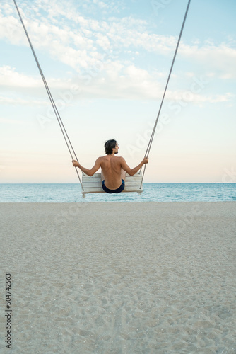 Young guy on rope swing on the beach on sea background. Back view. Vertical frame. Rest on the sea.