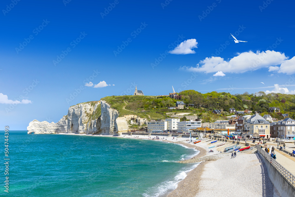 Panoramic view of the beach of Etretat in Normandy, a popular french seaside town known for its chalk cliffs, with Notre-Dame de la Garde chapel overlooking the Amont cliff on a sunny day