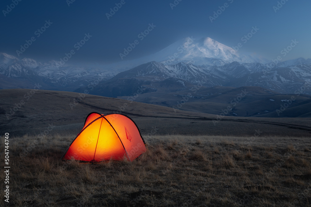Brightly lit tent at night in front of mountain ridge with large snow-covered Mount Elbrus, Russia