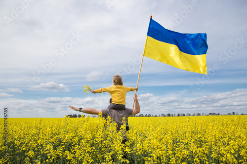 boy is sitting on shoulders of his father, holding large flag of Ukraine in hands, standing among yellow rapeseed field. education of patriotism. Pride, freedom. Ukrainians against war photo