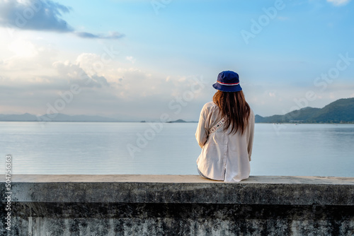 Back view of a female tourist looking away at river and mountain. Woman traveler sitting on concrete of lake or dam with a sunset background on a vacation trip. Asian lady enjoying nature on holiday.