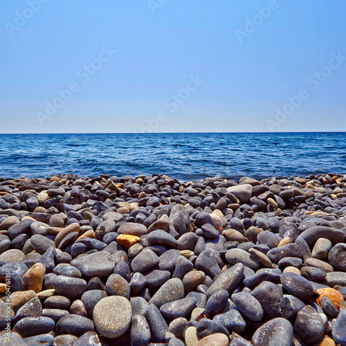 A black pebbles beach with blue sea and sky. Space for your text and logo.