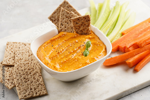 Paprika bell-pepper hummus dip looking like orange pumpkin puree, thin crackers with sesame seeds and carrot and cucumber cut in long strips and sticks on marble board photo