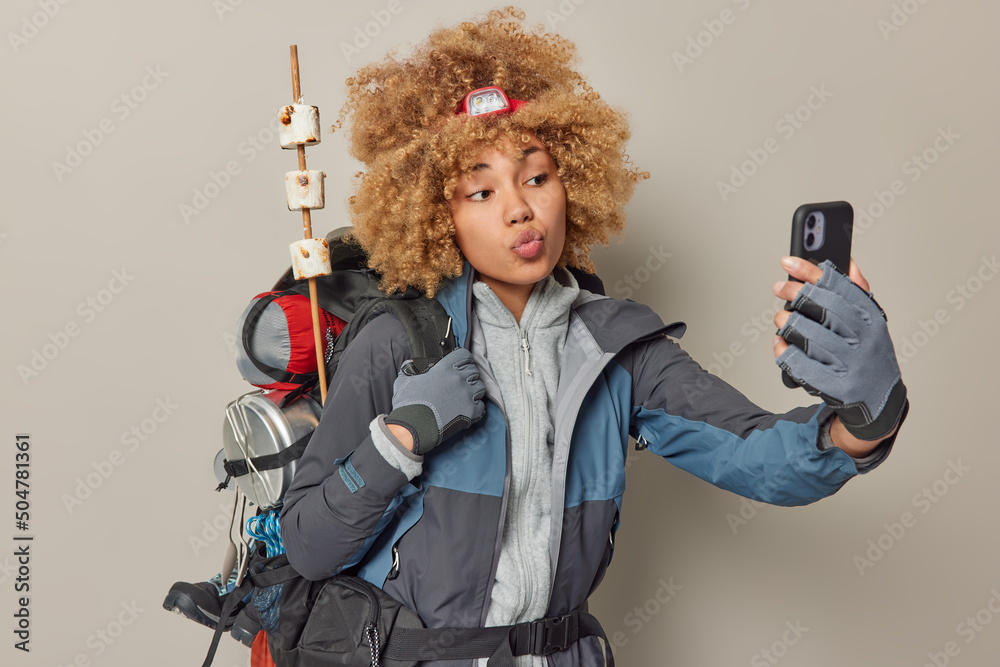 Active tourist takes selfie photo keeps lips folded dressed in sportswear carries heavy rucksack leads active lifestyle has journey expedition isolated over grey wall. Female hiker returns from picnic - obrazy, fototapety, plakaty 