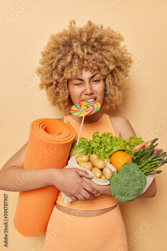 Vertical shot of curly woman breaks diet bires multicolored candy carries healthy vegetables grown in greenhouse goes in for sport regularly carries fitness mat poses indoor dressed in sportswear photo