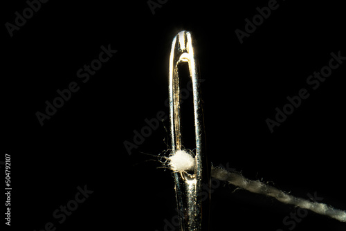 Macro image of a needle eye threaded with piece of white thread on black background. Steel shiny tailor's needle with permeation thread close up. The tool for sewing, embroidery, repair of clothes.