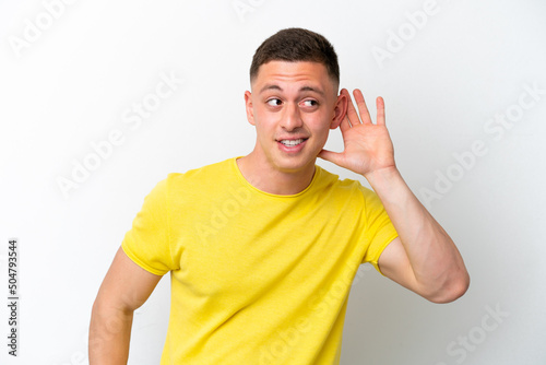 Young brazilian man isolated on white background listening to something by putting hand on the ear