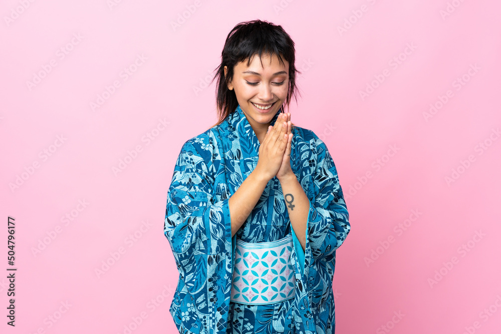 Young woman wearing kimono over isolated blue background keeps palm together. Person asks for something Young woman wearing kimono over isolated blue background keeps palm together. Person asks for something