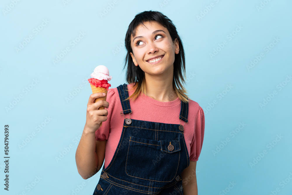 Young Uruguayan girl holding a cornet ice cream over isolated blue background looking up while smiling Young Uruguayan girl holding a cornet ice cream over isolated blue background looking up while smiling