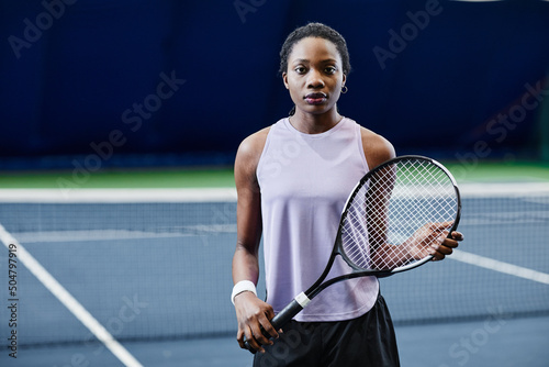 Waist up portrait of African American young woman holding tennis racket and looking at camera while posing confidently at indoor court, copy space © Seventyfour