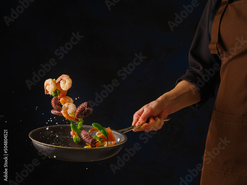 Fototapeta Naklejka Na Ścianę i Meble -  Seafood with vegetables and herbs in a frying pan on a black background in the chef's hand. Levitation. Sea Asian cuisine. Restaurant, hotel, picnic, banquet, recipe book.