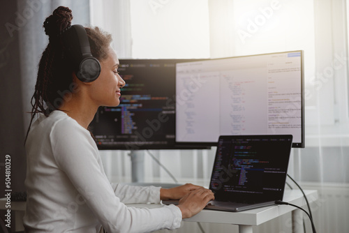 Cheerful beautiful female IT developer listening to music in headphones while coding at home