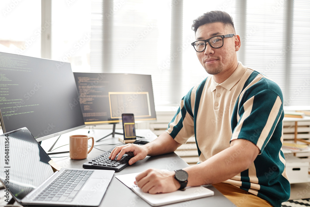 Warm toned portrait of Asian man as IT developer or QA engineer smiling at camera while sitting at workplace with computers, copy space Warm toned portrait of Asian man as IT developer or QA engineer smiling at camera while sitting at workplace with computers, copy space