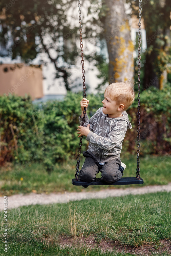 Little boy having fun on a swing on the playground in public park on sunny summer day. Happy child enjoy swinging. Active outdoors leisure for child in city.