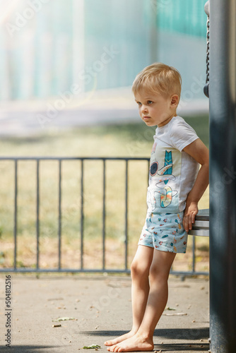 A blond, barefoot boy in a white shirt takes a break on an outdoor playground on a hot summer day