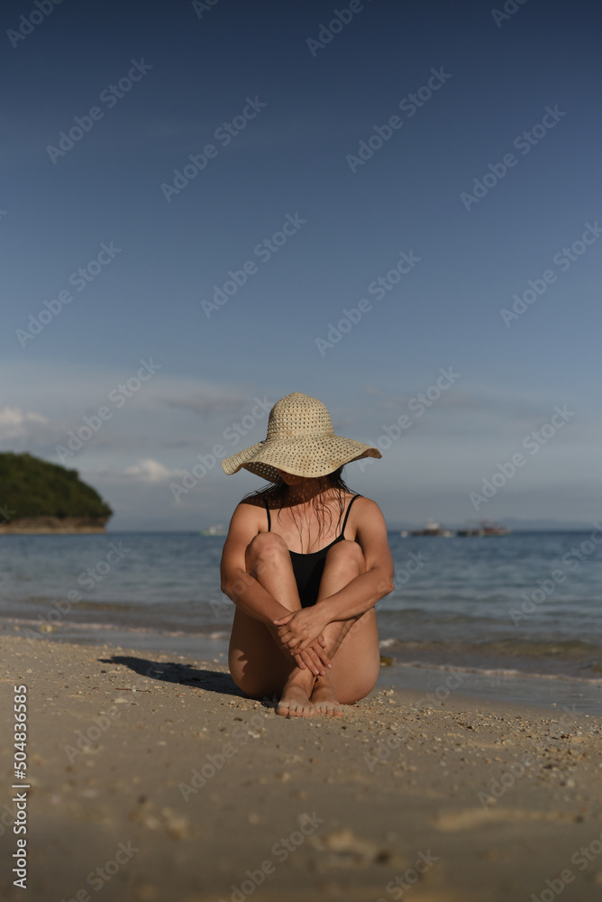 An elegant woman with hat is sitting on the beach. She is wearing a black swimsuit. Front view. Sea landscape, blue sky. Travel, vacation, holidays, tourism. Sea landscape, blue sky. Philippines