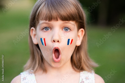Portrait of surprised 7 years old child with painted cheeks in flag of France walking outdoor. Cute little girl screaming with opened mouth and crazy expression looking at camera. 