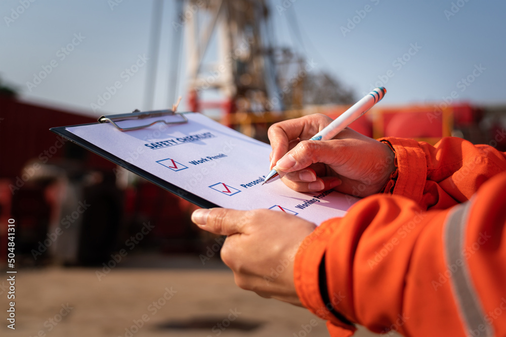 Construction worker is using a pen to checking on checklist - Safety audit and inspection working action with blurred background of machinery. Selective focus at hand. - obrazy, fototapety, plakaty 