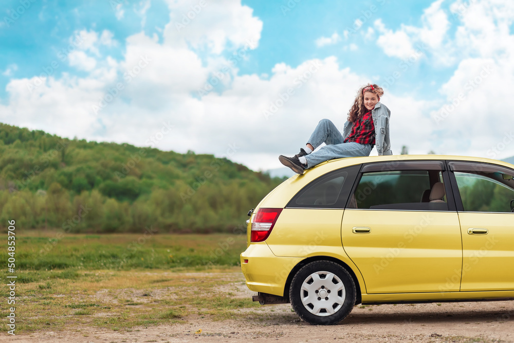 Pretty teen girl is sitting on the roof of a yellow car. Mountains and sky are in the background. Copy space. The concept of freedom and travel Pretty teen girl is sitting on the roof of a yellow car. Mountains and sky are in the background. Copy space. The concept of freedom and travel