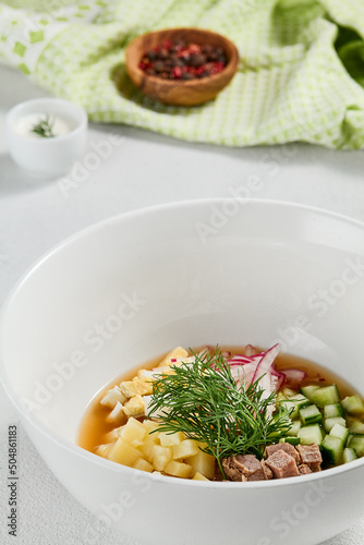 Traditional russian soup - okroshka with fresh vegetables, meat and kvass. Cold summer soup on white table. Aesthetic composition with traditional russian food and spices, textile, spoon. photo