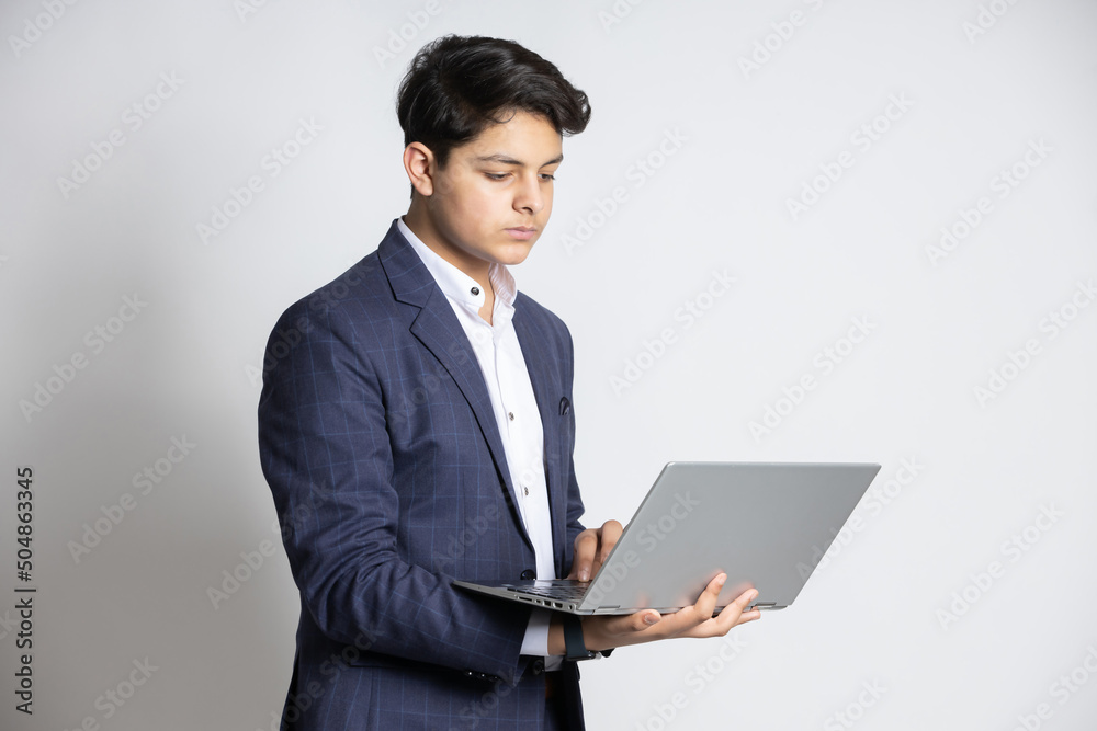 Young handsome smiling Indian teenager boy wearing suit working laptop at isolated on white studio background. Asian corporate male using computer. Young handsome smiling Indian teenager boy wearing suit working laptop at isolated on white studio background. Asian corporate male using computer.