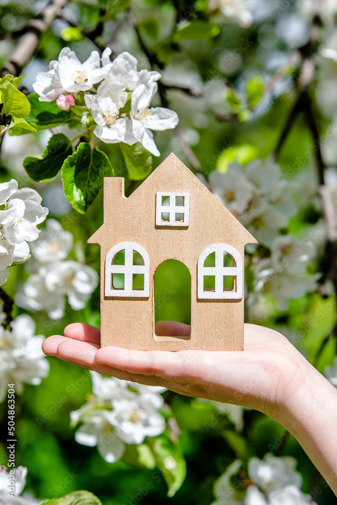 The girl holds the house symbol against the background of blossoming appletree
