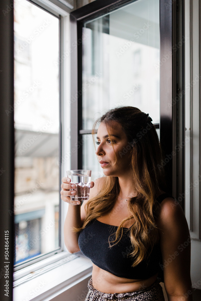 Young woman holding glass of water looking through window at home