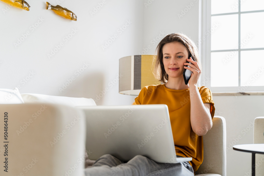Smiling freelancer talking on mobile phone sitting with laptop on sofa in living room