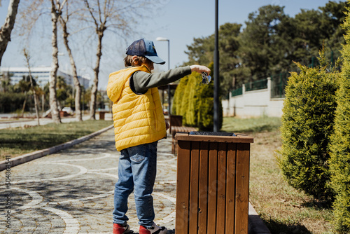 Schoolboy kid throwing the trash into dumpster. Boy using recycling bin to throw away the litter. Caucasian child recycles the junk into the trash-can.. photo