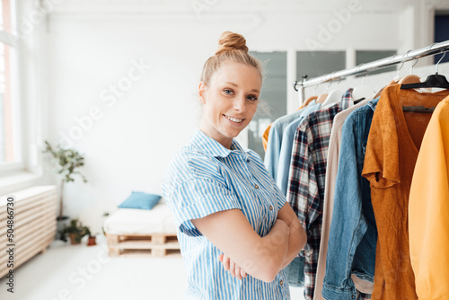 Smiling fashion designer with arms crossed standing near clothes rack on studio photo