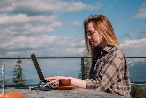 A young, beautiful girl in a plaid shirt talks on a smartphone sitting at an old wooden table in front of a laptop in nature and drinks coffee