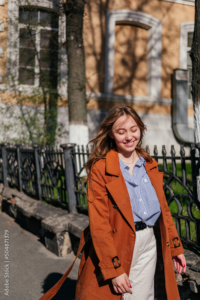 Portrait of smiling young woman in coat with closed eyes outdoors. Girl in city enjoying sun