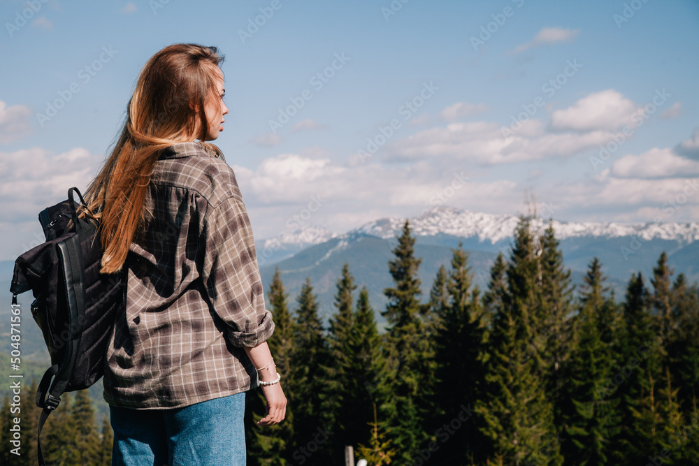 A young, slender girl with loose hair in a plaid shirt and jeans with a black backpack stands against the backdrop of mountains in sunny weather. View from the back.