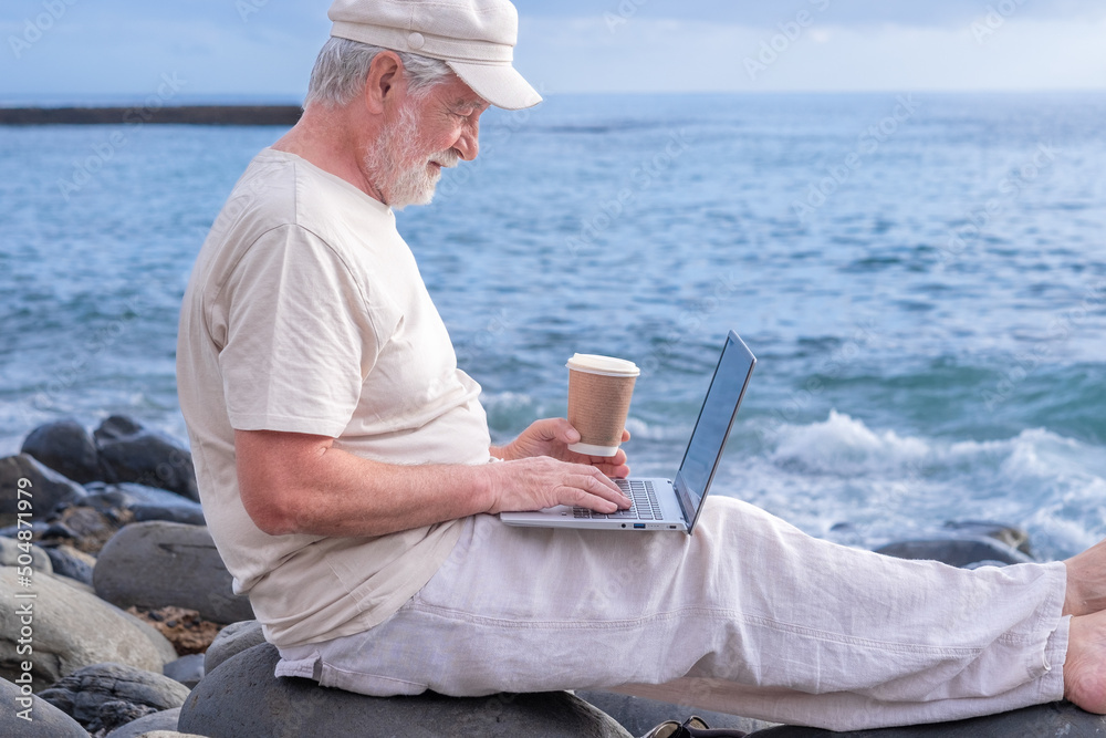 Senior active man sitting on the beach barefoot using laptop. Elderly bearded male holding a coffee cup enjoying sunset at sea, horizon over water
