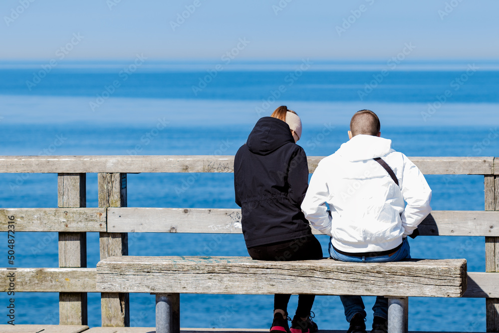 Young Couple Caucasians In Light Dark Clothes Sitting On Bench Opposite The Water Surface. View From The Back. Calm Blue Sea.