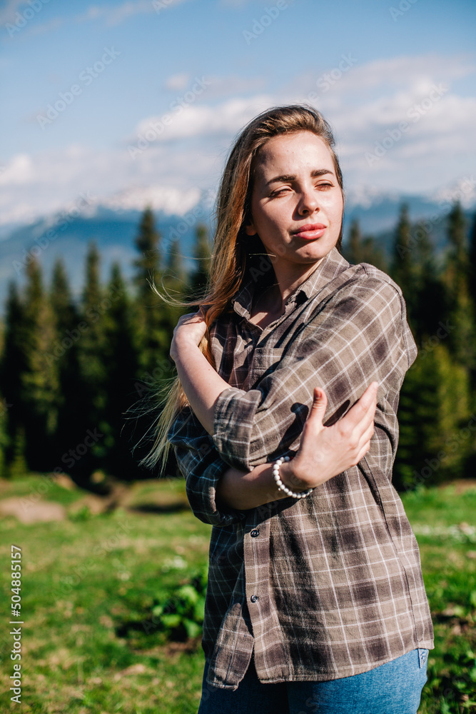 a young, slender girl with loose hair in a plaid shirt and jeans poses for sunny weather in the mountains