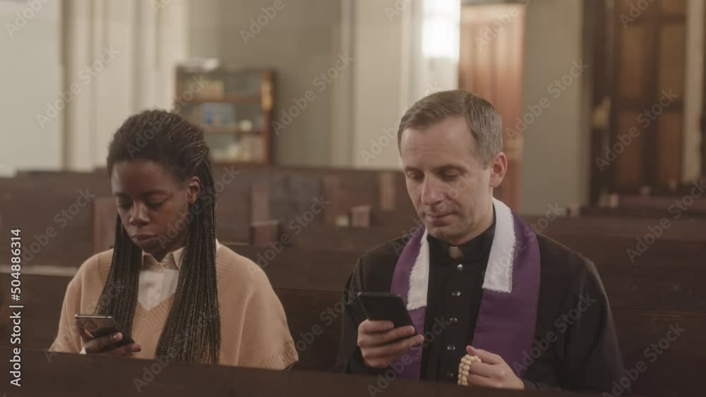 Slowmo of multiethnic priest and young female parishioner sitting next to each other on wooden bench in Catholic church using their smartphones Slowmo of multiethnic priest and young female parishioner sitting next to each other on wooden bench in Catholic church using their smartphones