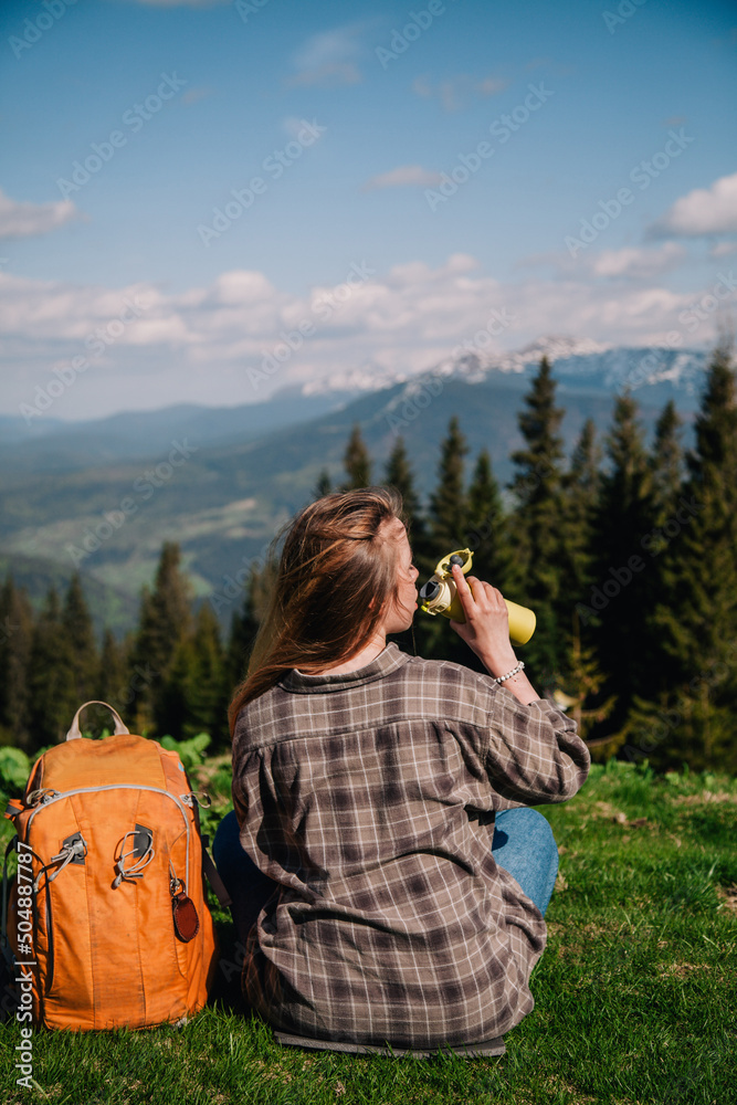 A young, slender girl with loose hair in a plaid shirt and jeans with an orange backpack drinks tea from a thermal mug sitting on the green grass against the backdrop of the mountains.