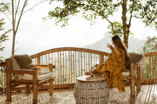 A girl is relaxing and drinking coffee on a balcony in a bamboo house overlooking the mountains. Coffee overlooking the mountains in Bali. © Yevhenii