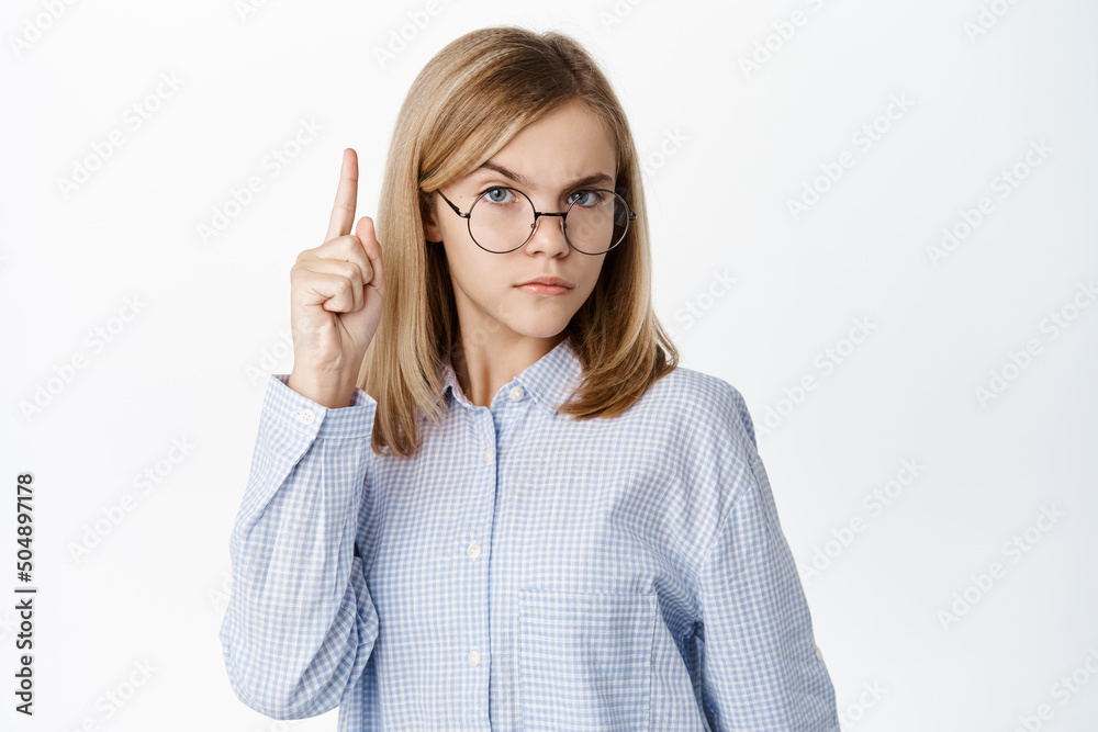 Smart cute little kid, girl in glasses raising one finger up, looking thoughtful and clever, standing over white background Smart cute little kid, girl in glasses raising one finger up, looking thoughtful and clever, standing over white background