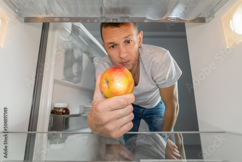 Man takes out a single apple from an empty refrigerator. Concept of delivery service, hunger, dients . Photo from inside the refrigerator