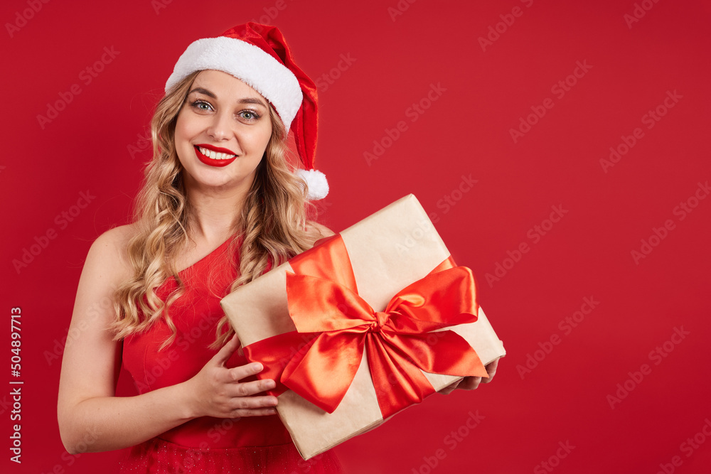 Pretty smiling lady in a red dress wearing a santa hat, holding a gift box with a bow. Pretty smiling lady in a red dress wearing a santa hat, holding a gift box with a bow.